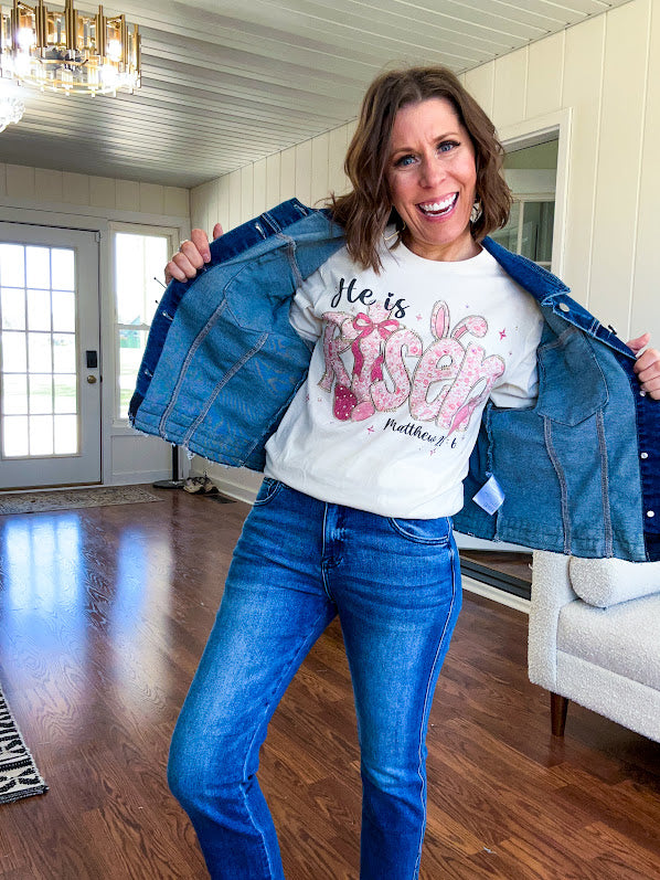 Cream cotton tee featuring "He is Risen" Matthew 28:6 in pink Easter graphics sprinkled with rhinestones.