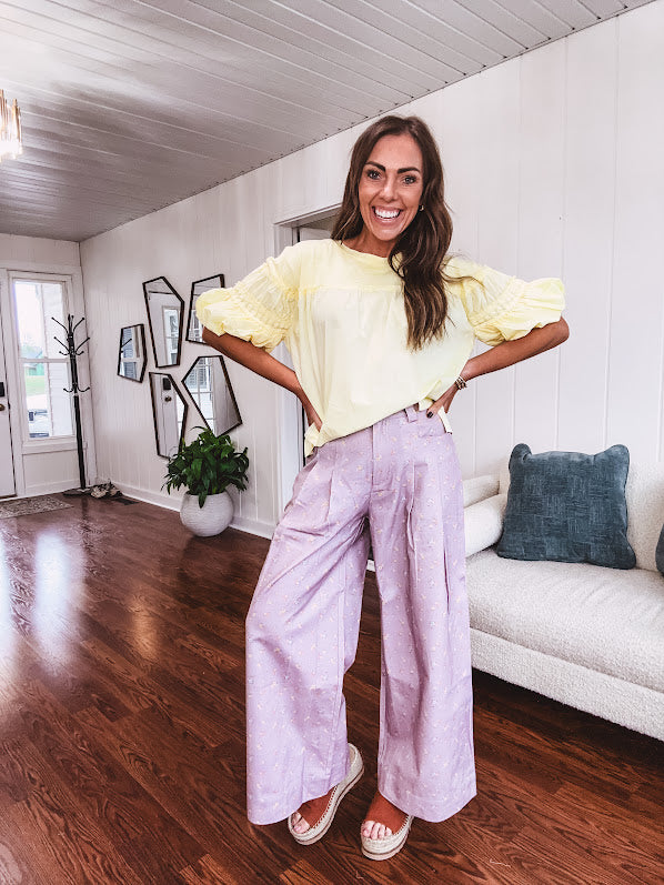 Light purple denim-like wide leg pants with elastic back waist, button and zipper closure and pleating at the front, with pockets and small pink and yellow florals throughout.