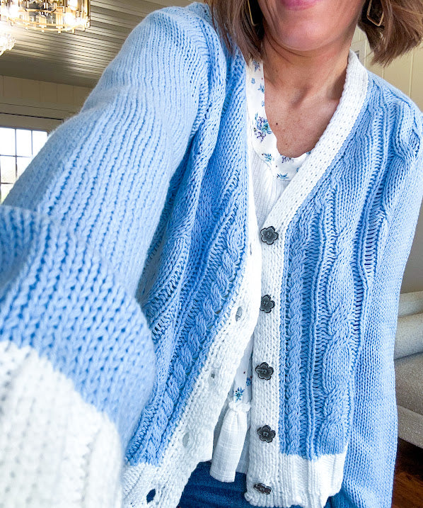 Light blue cable knit cardigan with white trim and flutter hem and cuffs with metal floral buttons down the front.