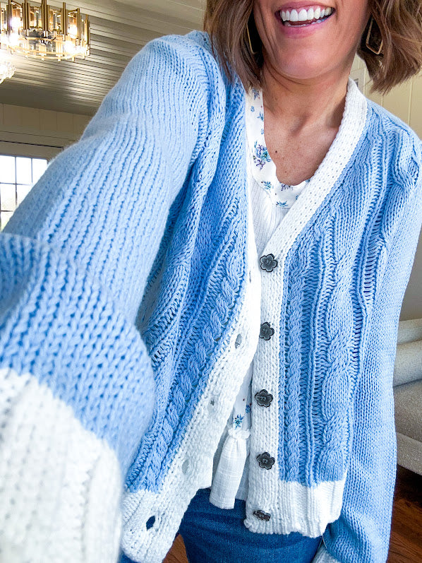 Light blue cable knit cardigan with white trim and flutter hem and cuffs with metal floral buttons down the front.