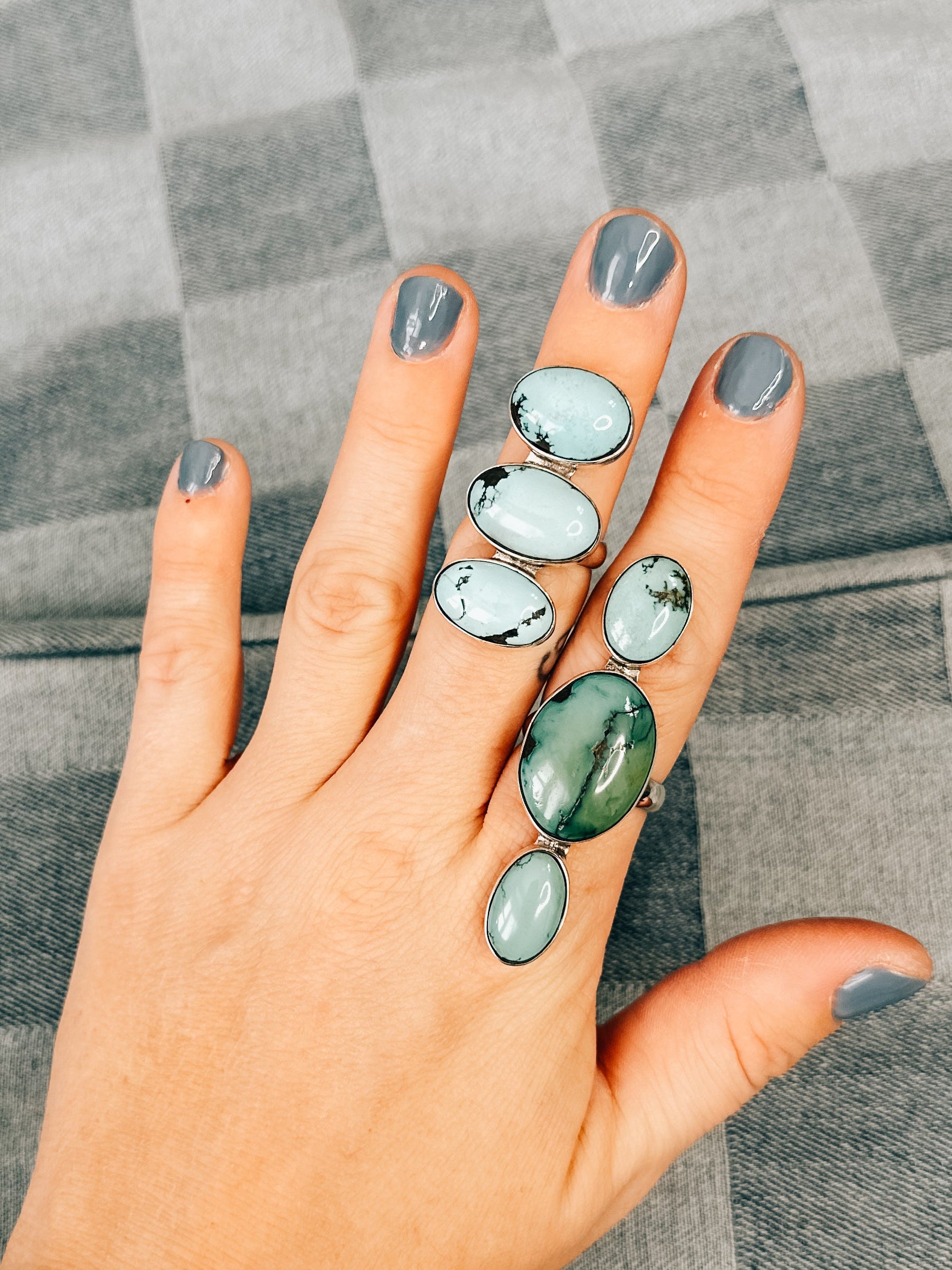 A hand displaying a triple ring with Kingman Turquoise stones, set in silver, on a textured grey background.