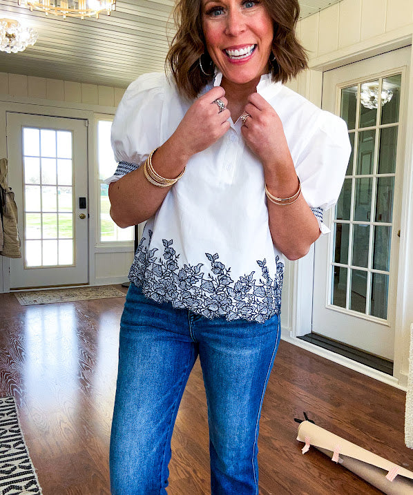 Crisp white blouse with ruffled mock neck and 4 button neckline, elastic puff sleeves and short hemline with navy floral embroidery at the hem and navy trim on the elastic at the sleeves.