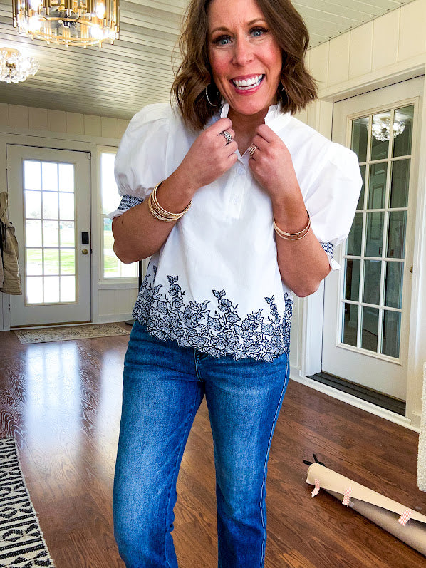 Crisp white blouse with ruffled mock neck and 4 button neckline, elastic puff sleeves and short hemline with navy floral embroidery at the hem and navy trim on the elastic at the sleeves.