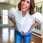 Crisp white blouse with ruffled mock neck and 4 button neckline, elastic puff sleeves and short hemline with navy floral embroidery at the hem and navy trim on the elastic at the sleeves.