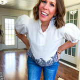 Crisp white blouse with ruffled mock neck and 4 button neckline, elastic puff sleeves and short hemline with navy floral embroidery at the hem and navy trim on the elastic at the sleeves.