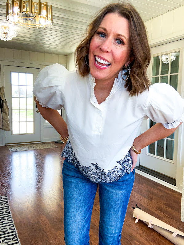 Crisp white blouse with ruffled mock neck and 4 button neckline, elastic puff sleeves and short hemline with navy floral embroidery at the hem and navy trim on the elastic at the sleeves.