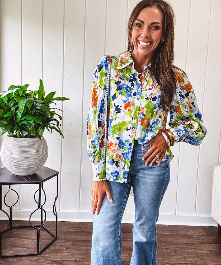 Bright green, orange and blue florals adorn this white button down blouse with a collar, puff sleeves and white eyelet trim throughout.