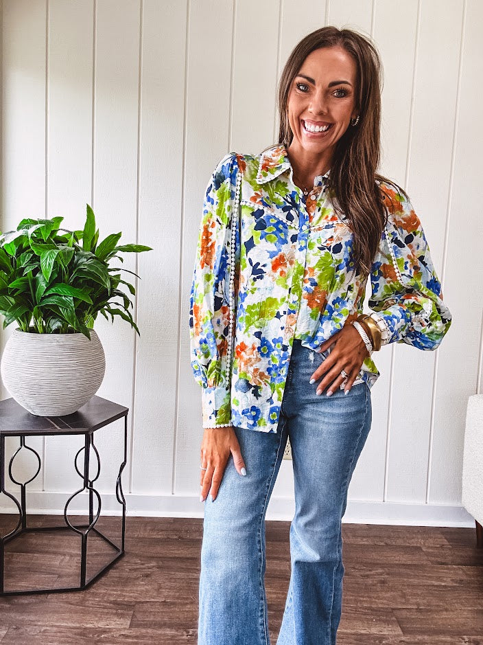 Bright green, orange and blue florals adorn this white button down blouse with a collar, puff sleeves and white eyelet trim throughout.