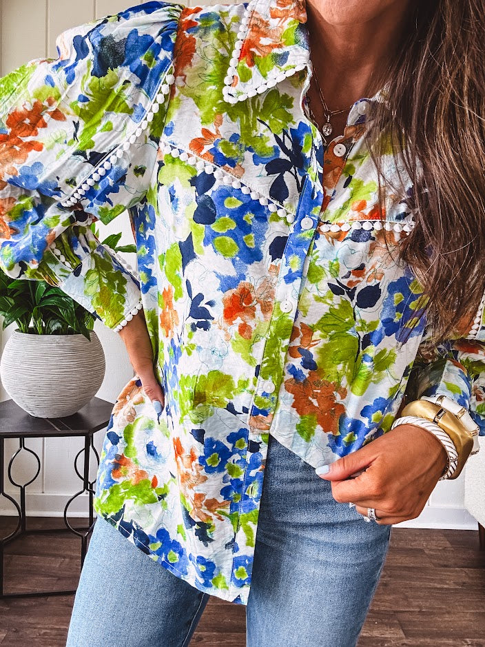 Bright green, orange and blue florals adorn this white button down blouse with a collar, puff sleeves and white eyelet trim throughout.