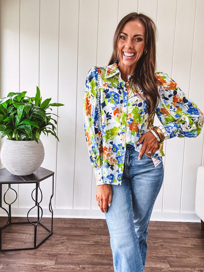 Bright green, orange and blue florals adorn this white button down blouse with a collar, puff sleeves and white eyelet trim throughout.