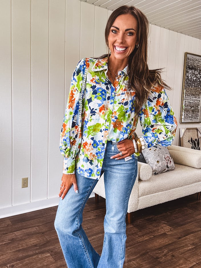 Bright green, orange and blue florals adorn this white button down blouse with a collar, puff sleeves and white eyelet trim throughout.