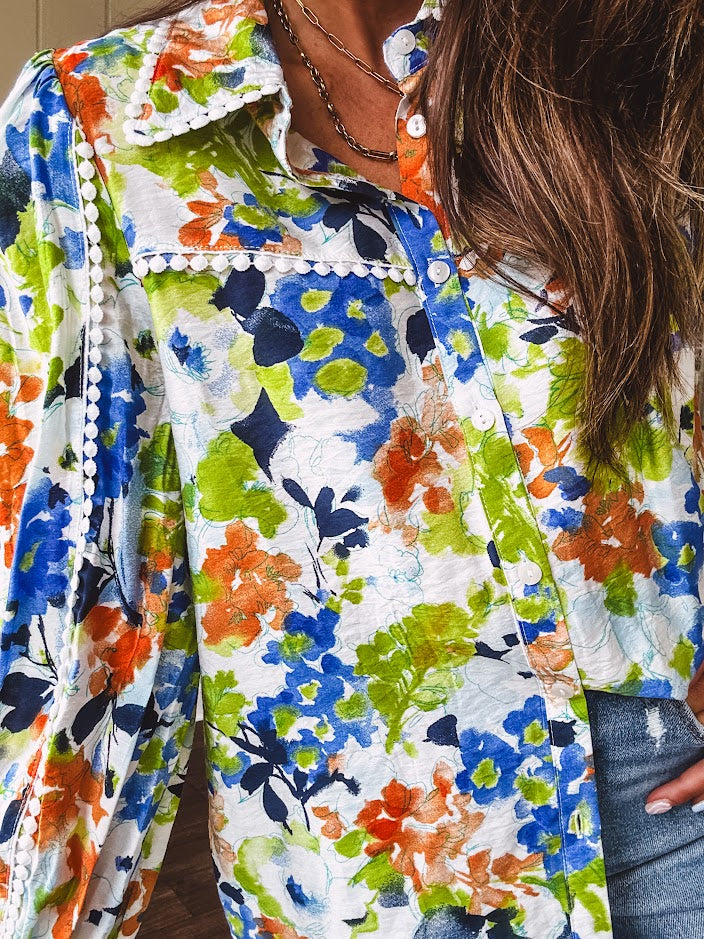 Bright green, orange and blue florals adorn this white button down blouse with a collar, puff sleeves and white eyelet trim throughout.