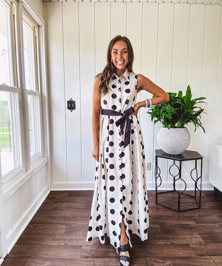 Maxi length ivory and black polka dot button front dress with collar, matching black waist tie and sleeveless design.