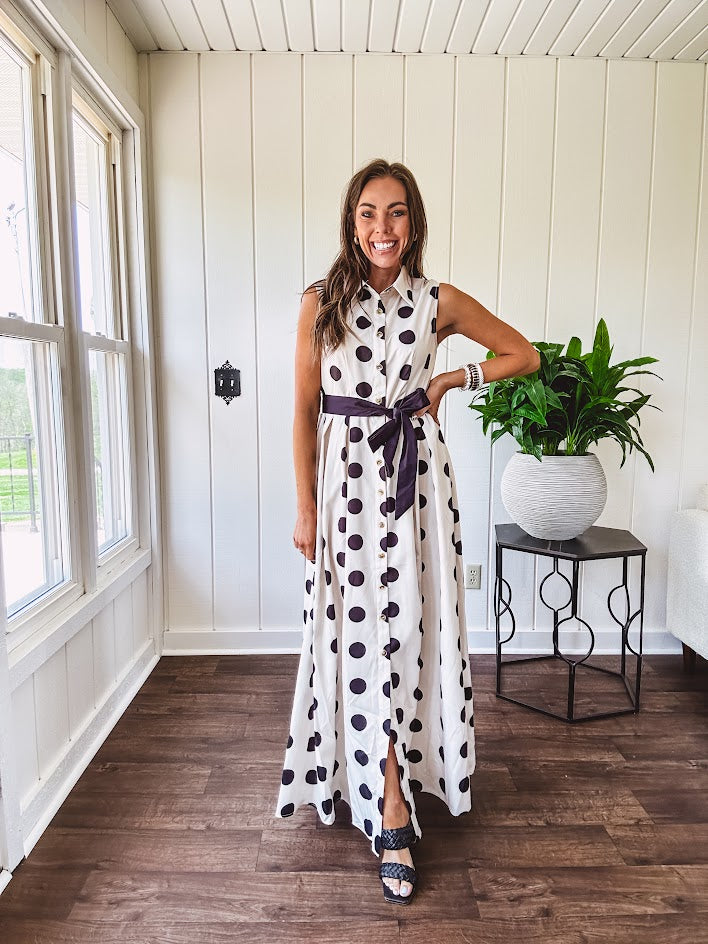 Maxi length ivory and black polka dot button front dress with collar, matching black waist tie and sleeveless design.
