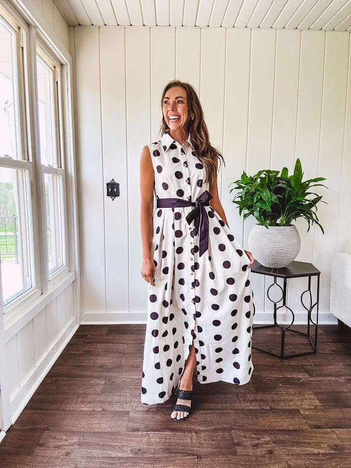 Maxi length ivory and black polka dot button front dress with collar, matching black waist tie and sleeveless design.