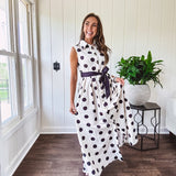 Maxi length ivory and black polka dot button front dress with collar, matching black waist tie and sleeveless design.