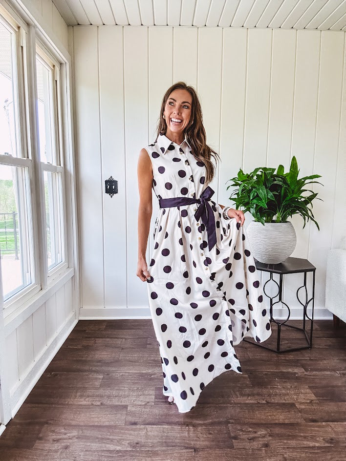 Maxi length ivory and black polka dot button front dress with collar, matching black waist tie and sleeveless design.