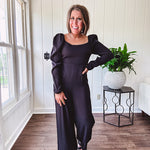 Black silky jumpsuit with puffed long sleeves, square neckline, side cutouts, elastic at the back waist and a tie at the back neckline.