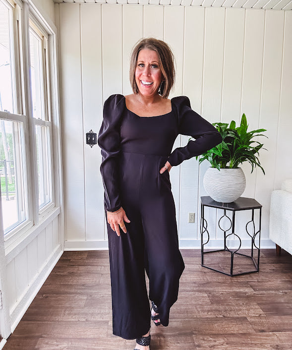 Black silky jumpsuit with puffed long sleeves, square neckline, side cutouts, elastic at the back waist and a tie at the back neckline.