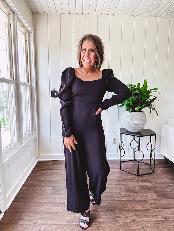 Black silky jumpsuit with puffed long sleeves, square neckline, side cutouts, elastic at the back waist and a tie at the back neckline.