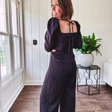 Black silky jumpsuit with puffed long sleeves, square neckline, side cutouts, elastic at the back waist and a tie at the back neckline.