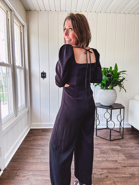 Black silky jumpsuit with puffed long sleeves, square neckline, side cutouts, elastic at the back waist and a tie at the back neckline.