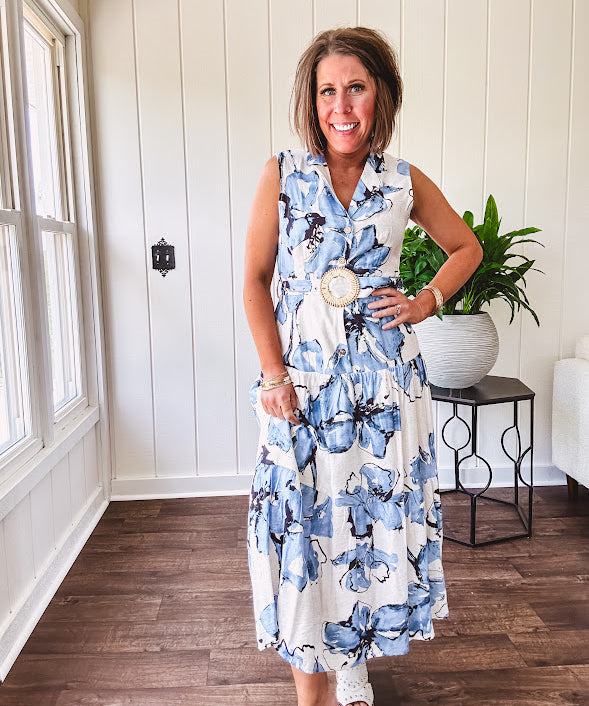 Lightweight cream sleeveless midi dress featuring blue and black large florals throughout, wooden buttons down to the lined tiered skirt, collar and matching belt.