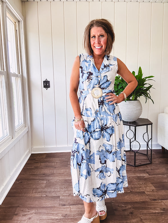 Lightweight cream sleeveless midi dress featuring blue and black large florals throughout, wooden buttons down to the lined tiered skirt, collar and matching belt.