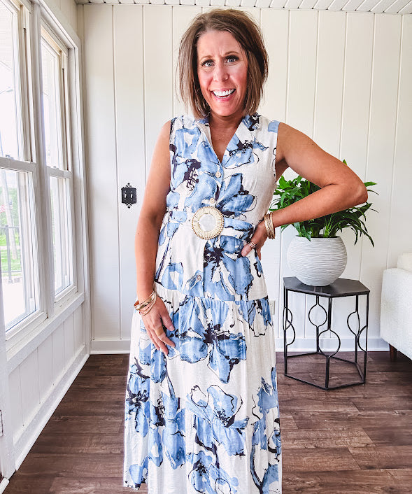 Lightweight cream sleeveless midi dress featuring blue and black large florals throughout, wooden buttons down to the lined tiered skirt, collar and matching belt.