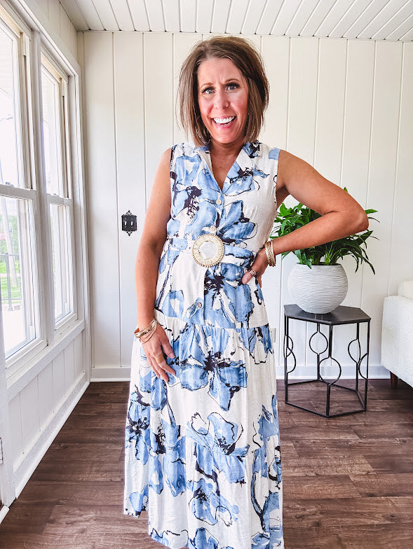 Lightweight cream sleeveless midi dress featuring blue and black large florals throughout, wooden buttons down to the lined tiered skirt, collar and matching belt.