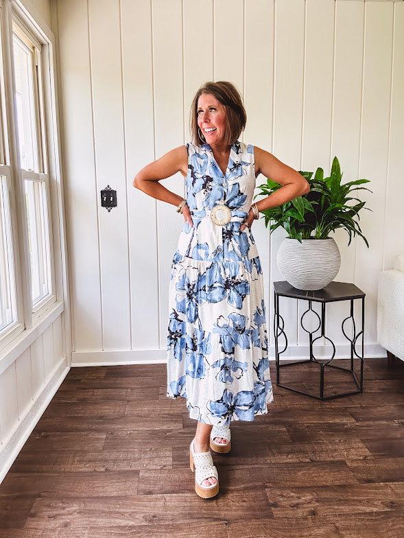 Lightweight cream sleeveless midi dress featuring blue and black large florals throughout, wooden buttons down to the lined tiered skirt, collar and matching belt.