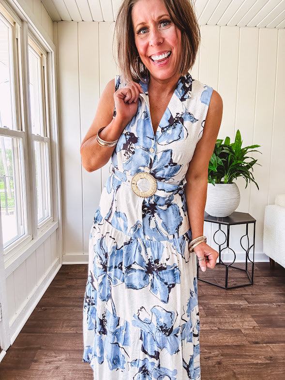 Lightweight cream sleeveless midi dress featuring blue and black large florals throughout, wooden buttons down to the lined tiered skirt, collar and matching belt.