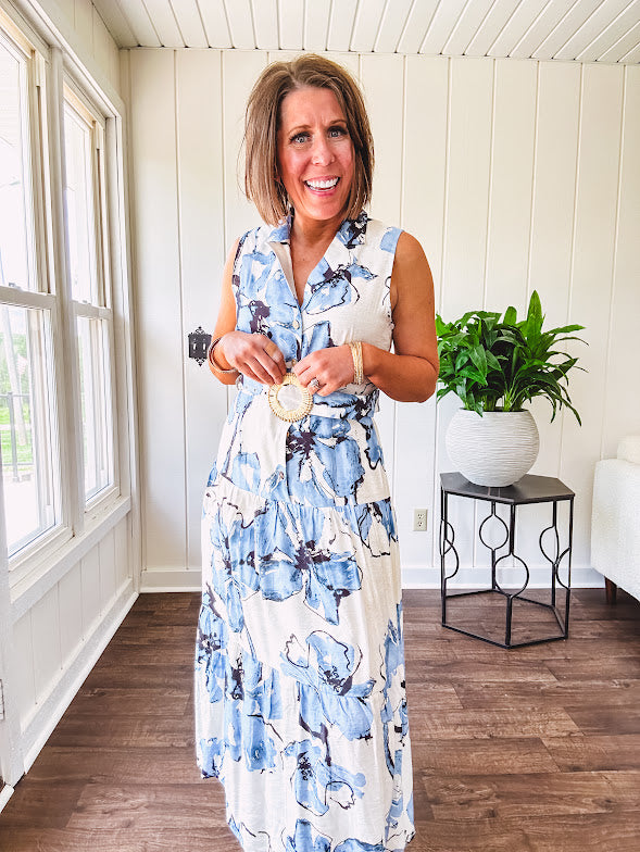 Lightweight cream sleeveless midi dress featuring blue and black large florals throughout, wooden buttons down to the lined tiered skirt, collar and matching belt.