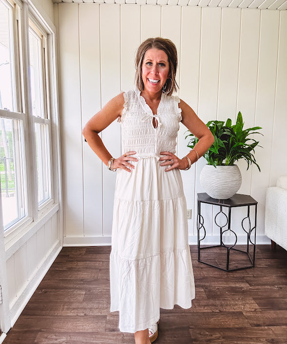 Oatmeal colored sleeveless midi dress with ruffled sleeves and collar with a tie, wide smocked bodice and lined, tiered skirt.