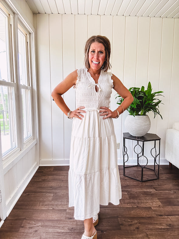 Oatmeal colored sleeveless midi dress with ruffled sleeves and collar with a tie, wide smocked bodice and lined, tiered skirt.