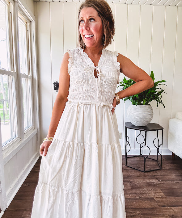 Oatmeal colored sleeveless midi dress with ruffled sleeves and collar with a tie, wide smocked bodice and lined, tiered skirt.