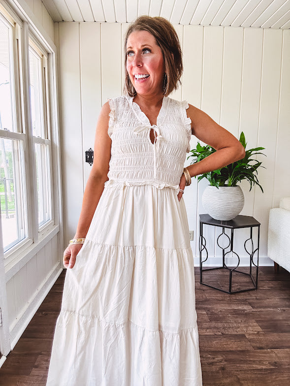 Oatmeal colored sleeveless midi dress with ruffled sleeves and collar with a tie, wide smocked bodice and lined, tiered skirt.