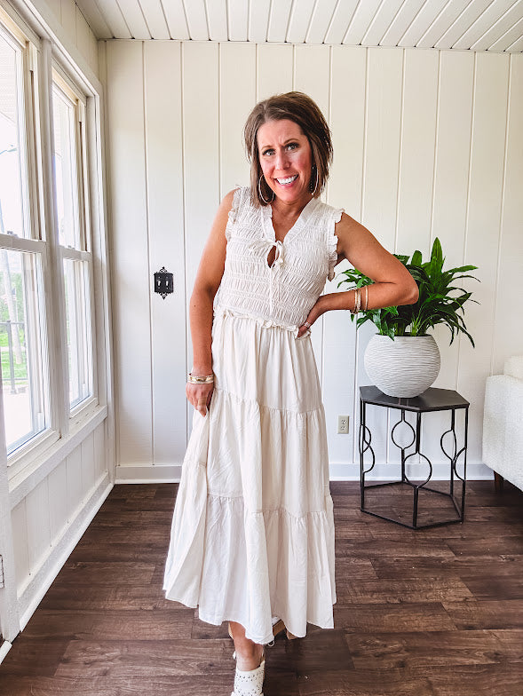 Oatmeal colored sleeveless midi dress with ruffled sleeves and collar with a tie, wide smocked bodice and lined, tiered skirt.