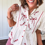 Thin, cotton linen blend cream top with short, puff sleeves, mock neck collar, red buttons down the front, a ruffle at the waist and red bow and floral embroidery throughout.