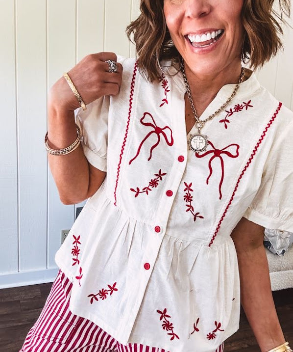 Thin, cotton linen blend cream top with short, puff sleeves, mock neck collar, red buttons down the front, a ruffle at the waist and red bow and floral embroidery throughout.