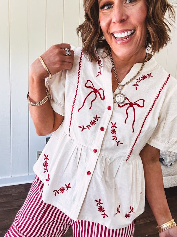 Thin, cotton linen blend cream top with short, puff sleeves, mock neck collar, red buttons down the front, a ruffle at the waist and red bow and floral embroidery throughout.