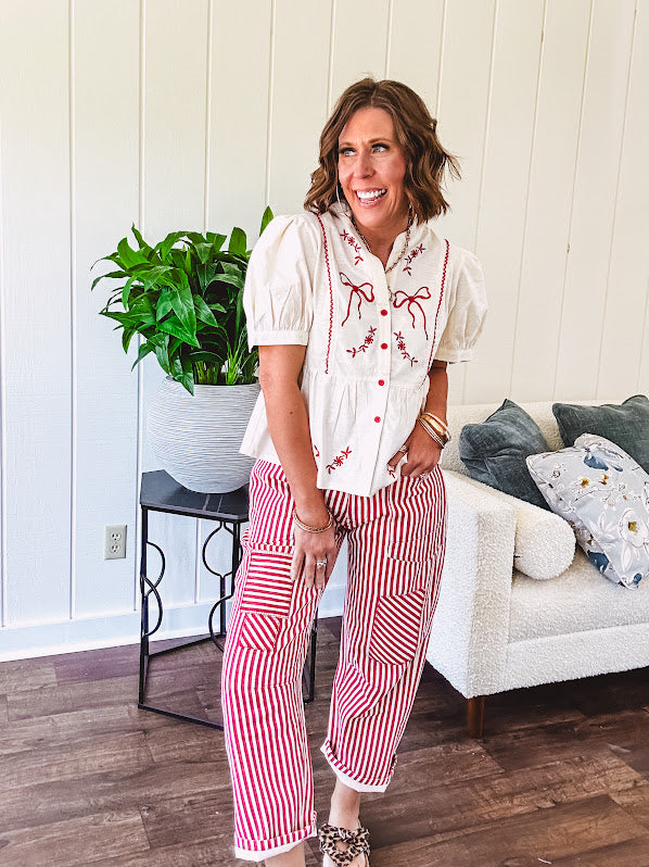 Thin, cotton linen blend cream top with short, puff sleeves, mock neck collar, red buttons down the front, a ruffle at the waist and red bow and floral embroidery throughout.