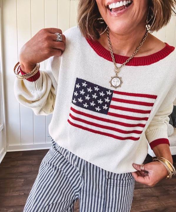 Cream long sleeved sweater with round neck and red trim at neck an cuffs with American Flag motif.