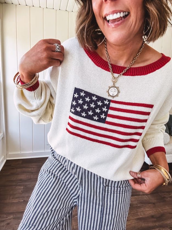 Cream long sleeved sweater with round neck and red trim at neck an cuffs with American Flag motif.