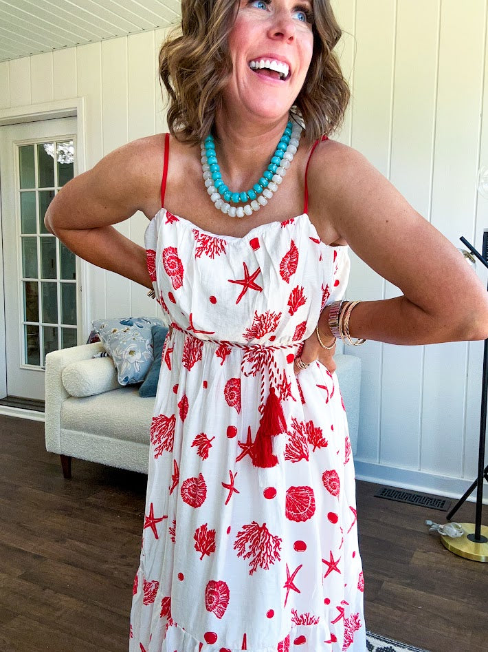 This fully lined white midi features an eye-catching red shell print that demands attention, paired with adjustable red spaghetti straps and a show-stopping red and white striped rope belt complete with charming tassels.