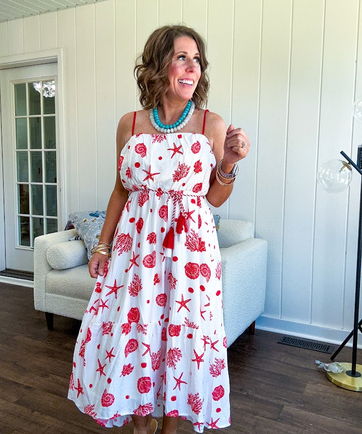 This fully lined white midi features an eye-catching red shell print that demands attention, paired with adjustable red spaghetti straps and a show-stopping red and white striped rope belt complete with charming tassels.