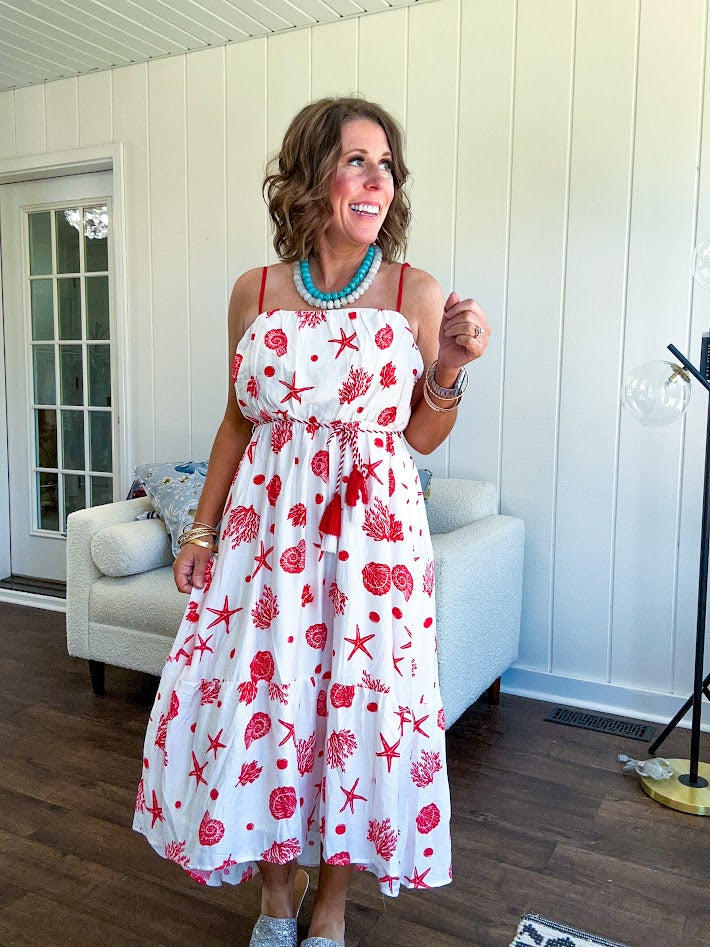 This fully lined white midi features an eye-catching red shell print that demands attention, paired with adjustable red spaghetti straps and a show-stopping red and white striped rope belt complete with charming tassels.