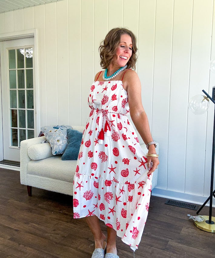 This fully lined white midi features an eye-catching red shell print that demands attention, paired with adjustable red spaghetti straps and a show-stopping red and white striped rope belt complete with charming tassels.