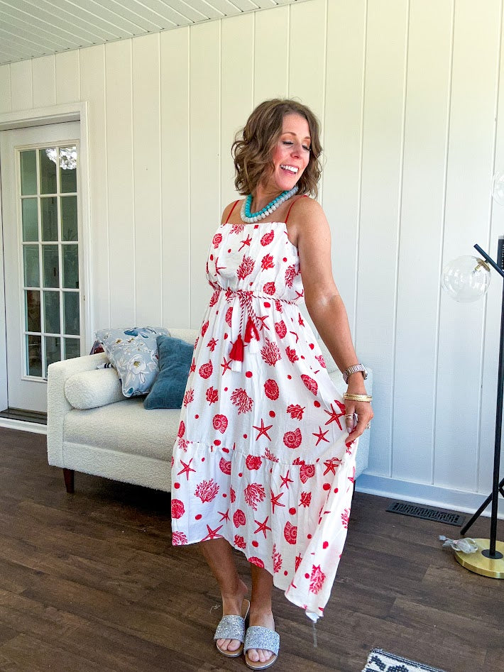 This fully lined white midi features an eye-catching red shell print that demands attention, paired with adjustable red spaghetti straps and a show-stopping red and white striped rope belt complete with charming tassels.