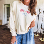Cropped creamy light weight crew pullover with long sleeves and American flag embroidery with a light blue bow on the front.